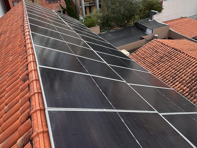 Sistema fotovoltaico instalado sobre telhado cerâmico de residência, com os telhados do bairro ao fundo